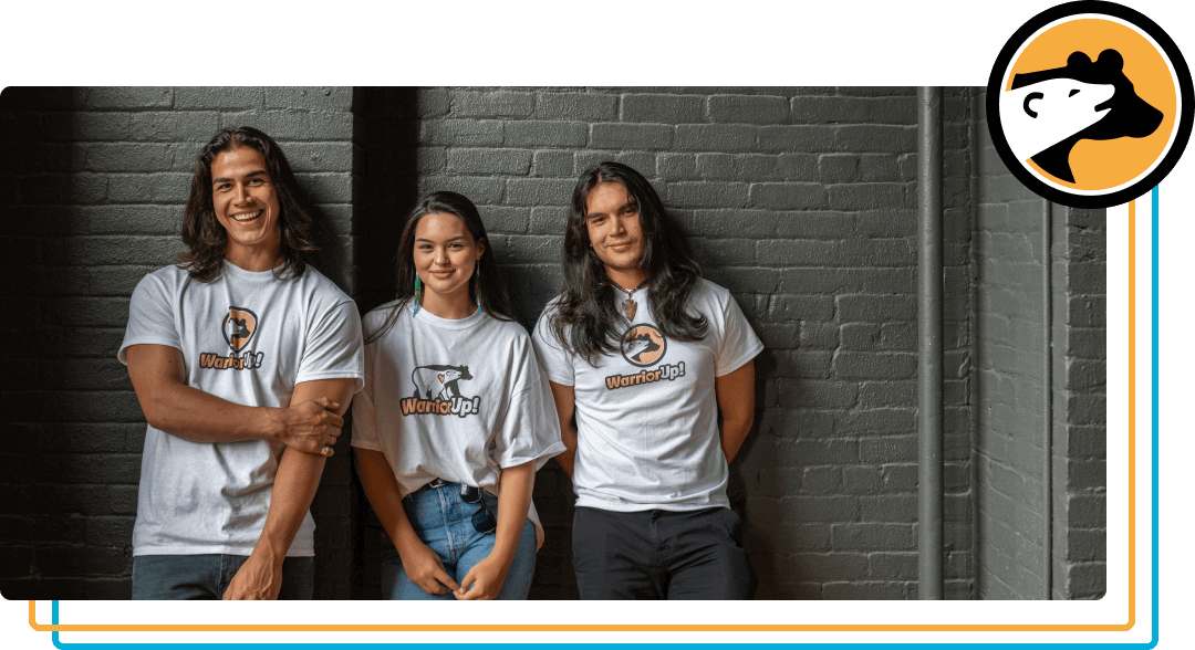 Three smiling young people stand against a dark grey brick wall. They are all of medium complexion with long hair. They all are wearing white t-shirts that say "Warrior Up!" on them.